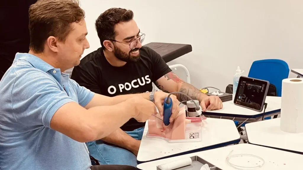 Treinamento de POCUS do EcoVerso: participante manuseia o transdutor sobre um phantom vascular, com aparelho portátil exibindo a imagem, acompanhado por instrutor